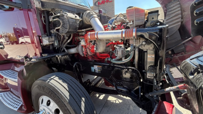 Kenworth T680 engine bay showing V-shaped diesel engine and exposed components, air intake tubing, hoses, and front suspension in view.