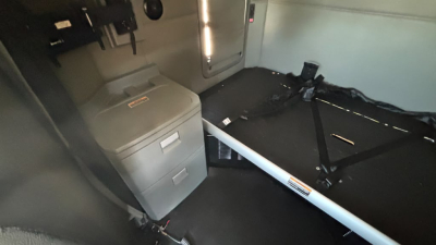 Vehicle interior showing sleeper area of a Kenworth T680 with a small filing cabinet, seating belt, and a storage area.