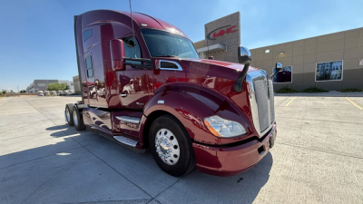 Kenworth T680 tractor-trailer front-left view with maroon paint and chrome wheels. Windshield shows no visible text. Background shows dealership building.