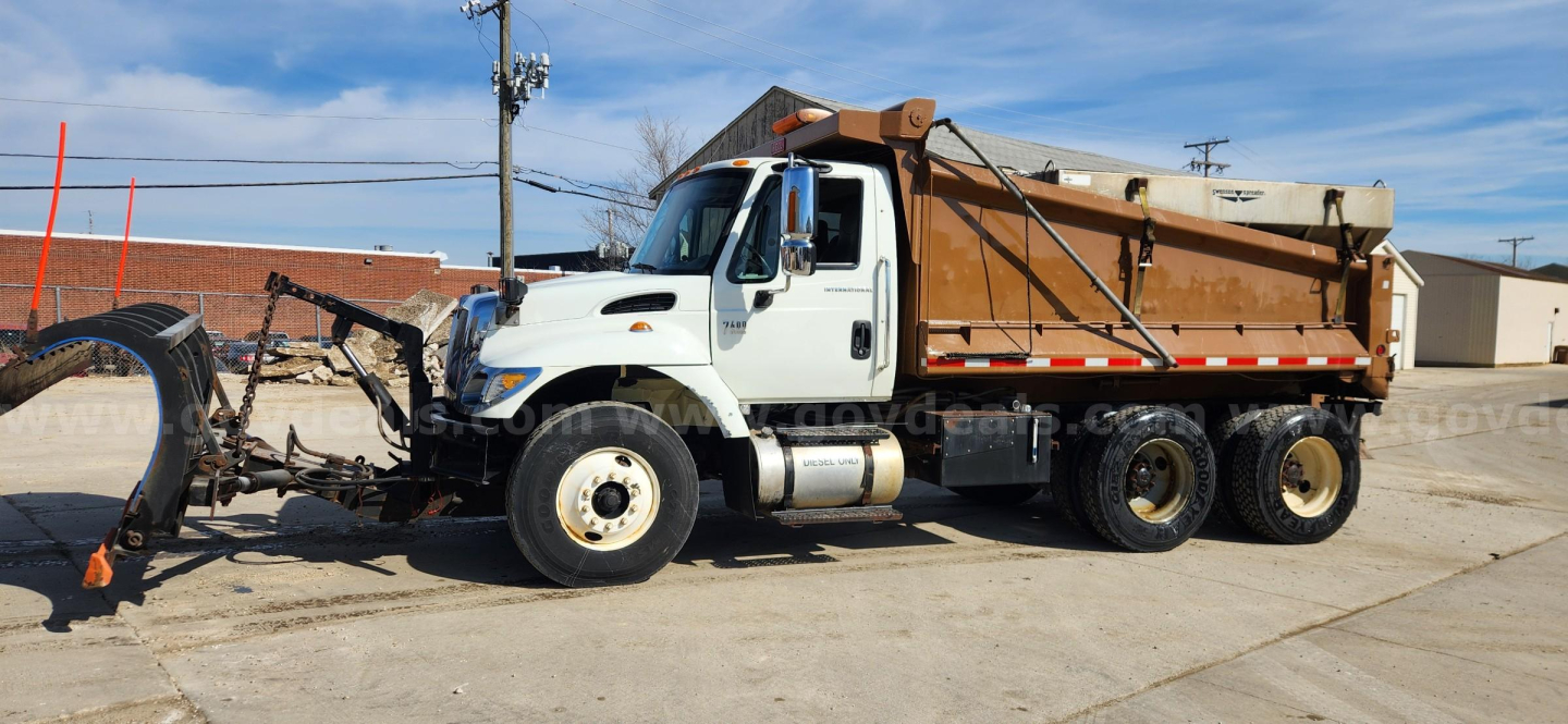 International 7400 dump truck with front-mounted plow and detached dump body, white cab, tan dump bed, side mirrors, and dual rear wheels in a lot.