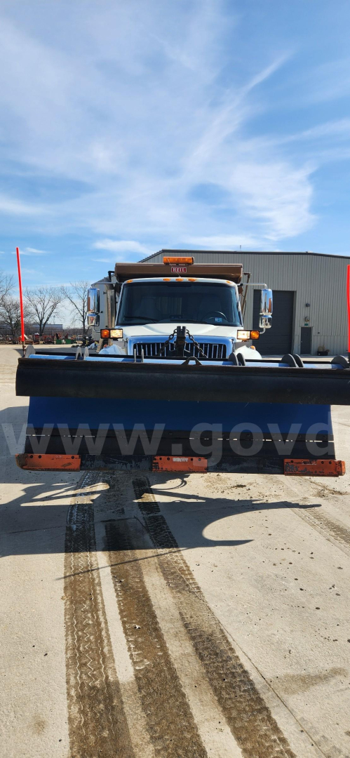 Front view of a white International dump truck equipped with a snow plow blade and amber warning lights on the cab, visible text includes a small yellow/amber sign above the windshield and the plow blade at the bottom.