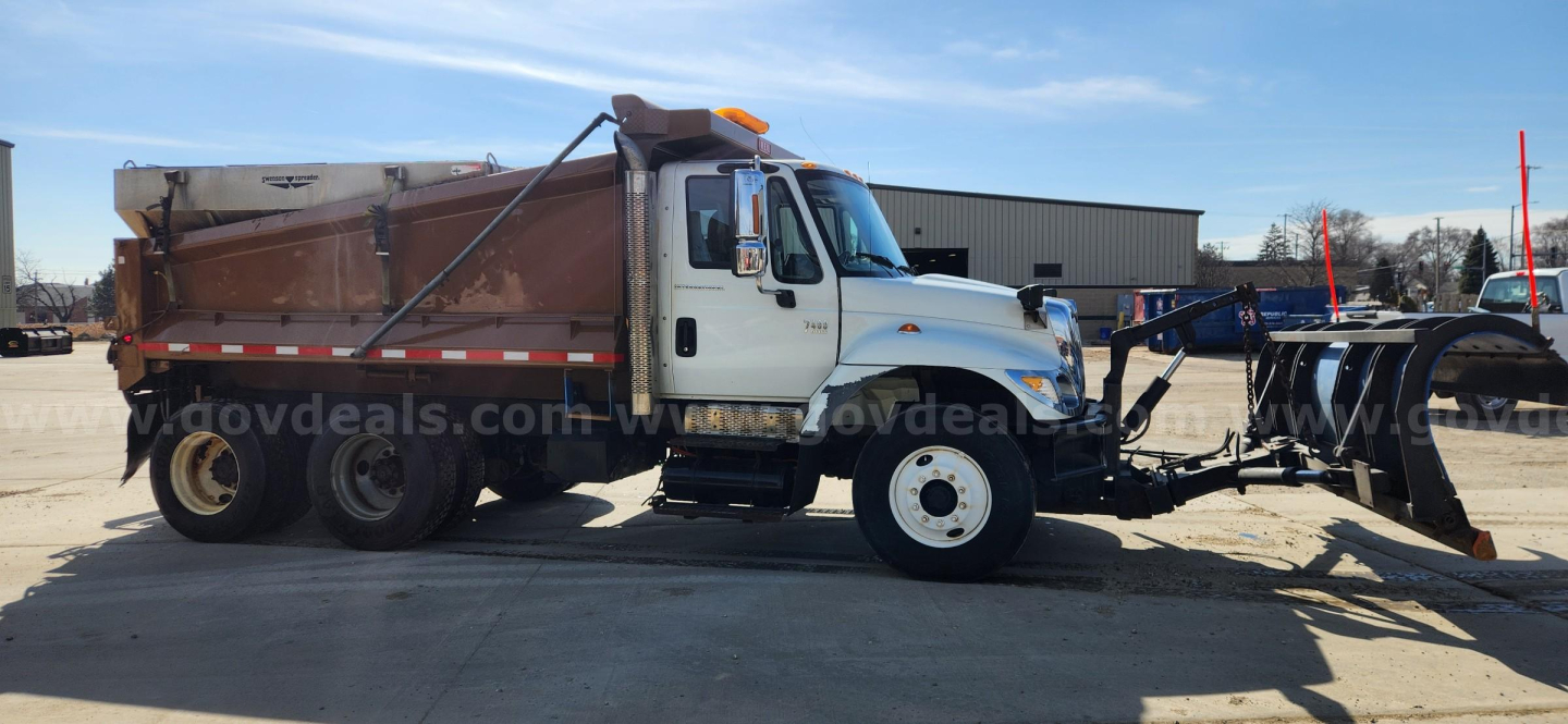 Dump truck with white cab and brown dump bed, side view showing large tires, a plow/mount in front, and a banner across the image with a vehicle marketplace watermark.