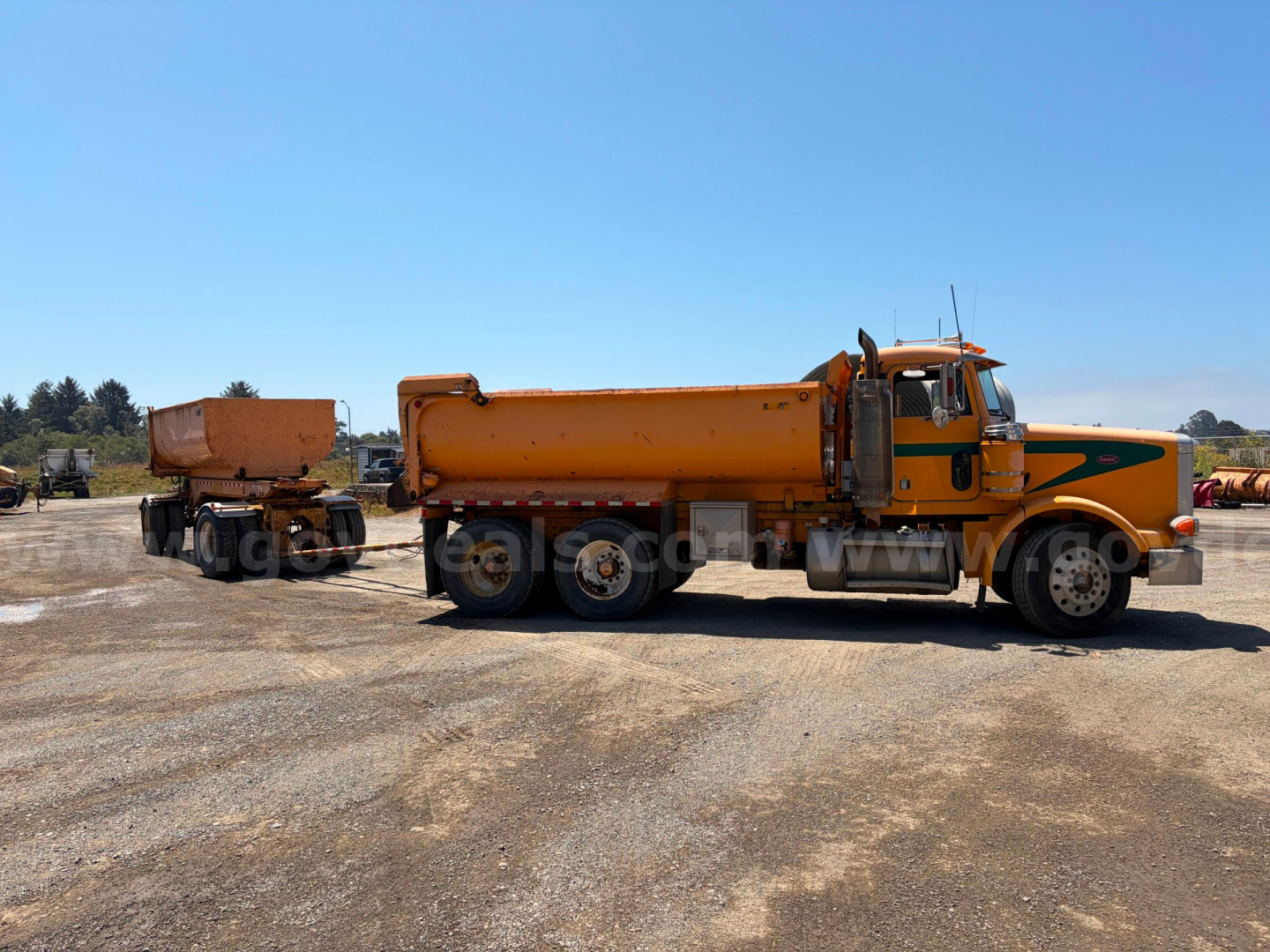 Peterbilt 378 dump truck tractor with orange cab and matching dump trailer connected, side view, clear sky and gravel lot.
