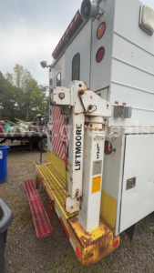 Truck rear showing Liftmoore hydraulic lift arm with L2? label and warning lights. Side of utility box with striped rear and steps. Visible text: LIFTMOORE on arm, tail lights, metal platform, rust patches.