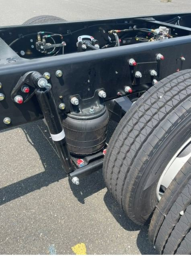 Chassis view of a new 2027 Hino L6 box van truck showing frame, air suspension canister, hydraulic lines, and dual rear tires.