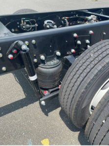Chassis view of a new 2027 Hino L6 box van truck showing frame, air suspension canister, hydraulic lines, and dual rear tires.