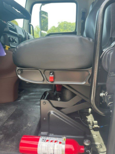 Truck interior seat area of a Hino L6 box van showing driver's seat, seat adjustment panel with red latch, and floor area near step.