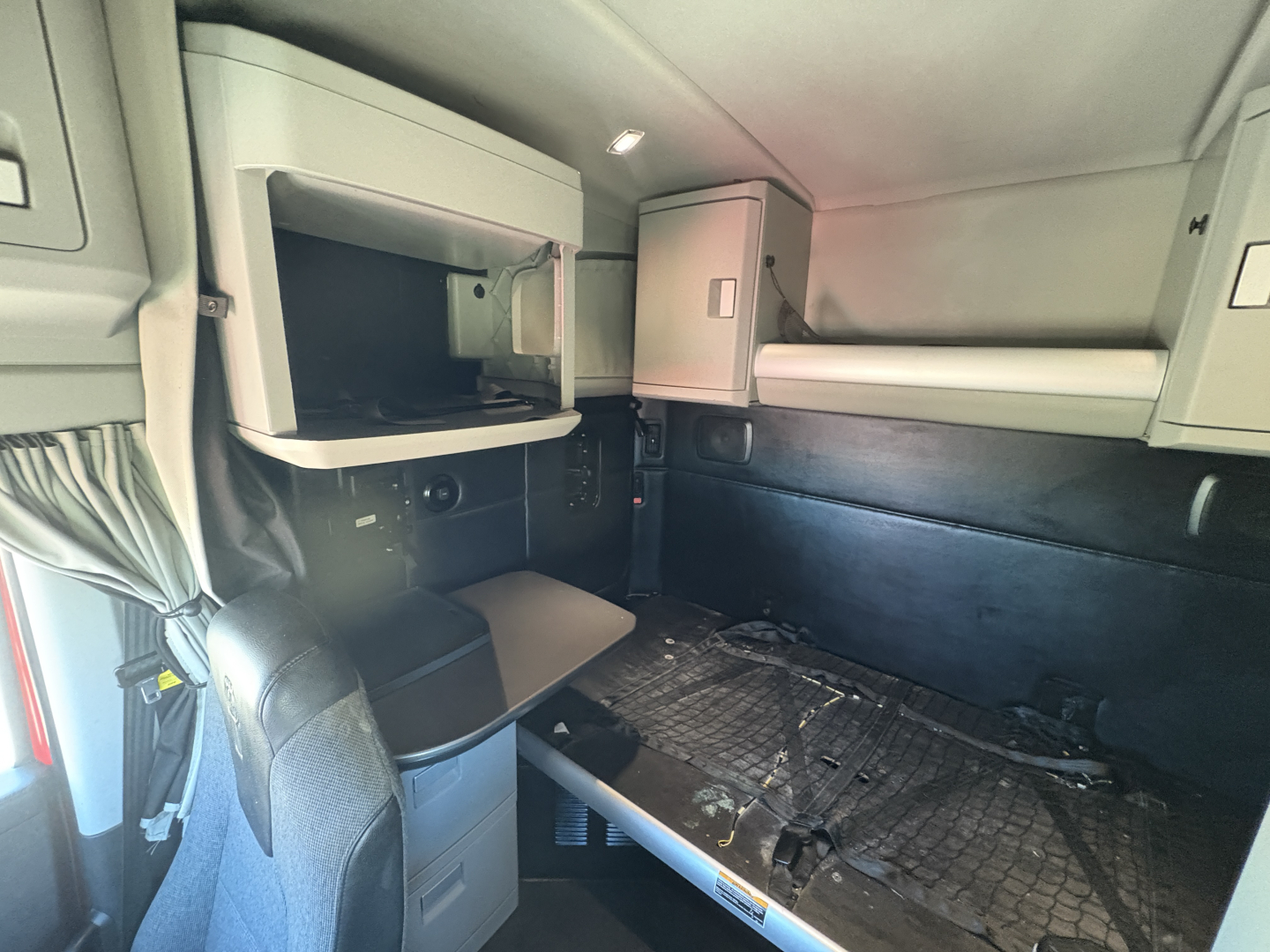 Kenworth T680 cabin interior showing sleeper cab with overhead storage, bunk area, and passenger seat; gray cabinets and window shade.