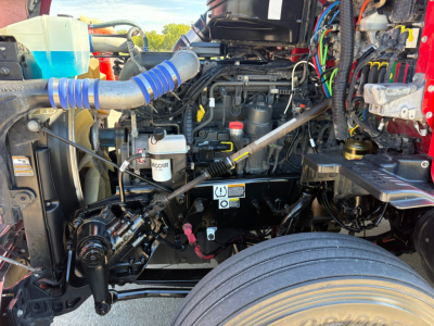 Kenworth truck engine bay with visible hoses, wiring, and battery area; tag labels and fluid reservoirs visible.