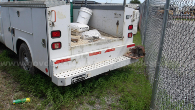 Ford F-250 service/utility truck rear view showing open cargo bed with equipment and a tow hitch, white utility body, chain-link fence in background.