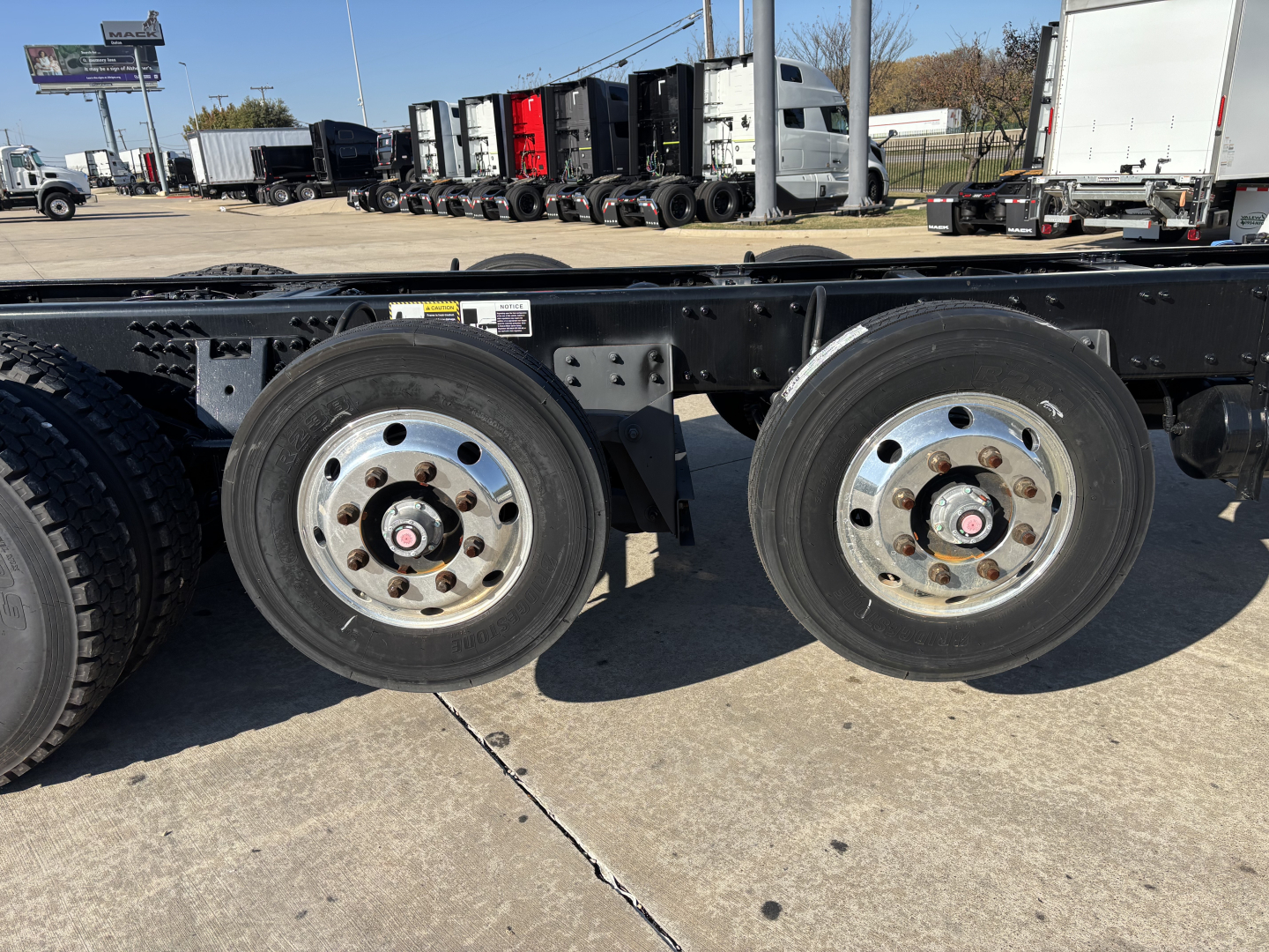 Dual rear axles on a Mack GR64B cab and chassis showing steel wheels and tires, framed undercarriage, and a row of trucks in the background.