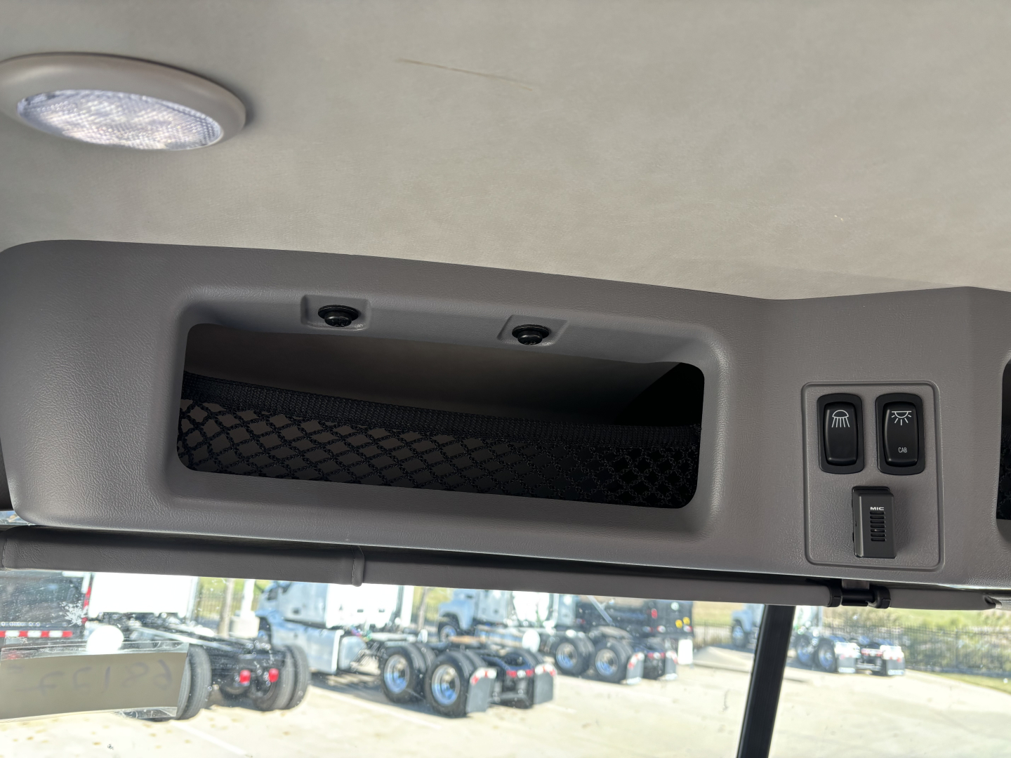 Mack GR64B cab interior upper dash area showing storage slot and control switches with window and exterior lot in background.