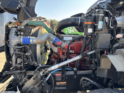 Engine bay of 2025 Mack GR64B cab and chassis showing coolant hose, red engine block, black plastic ducts, battery and wiring harness, various labels, and metal frame components.