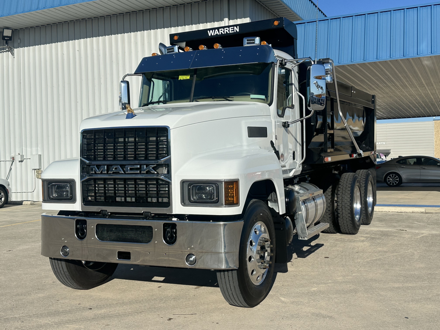 Mack PI64 dump truck in white cab with black dump bed, front grille with MACK emblem, large chrome bumper and dual rear wheels, factory wheels, showroom lot.