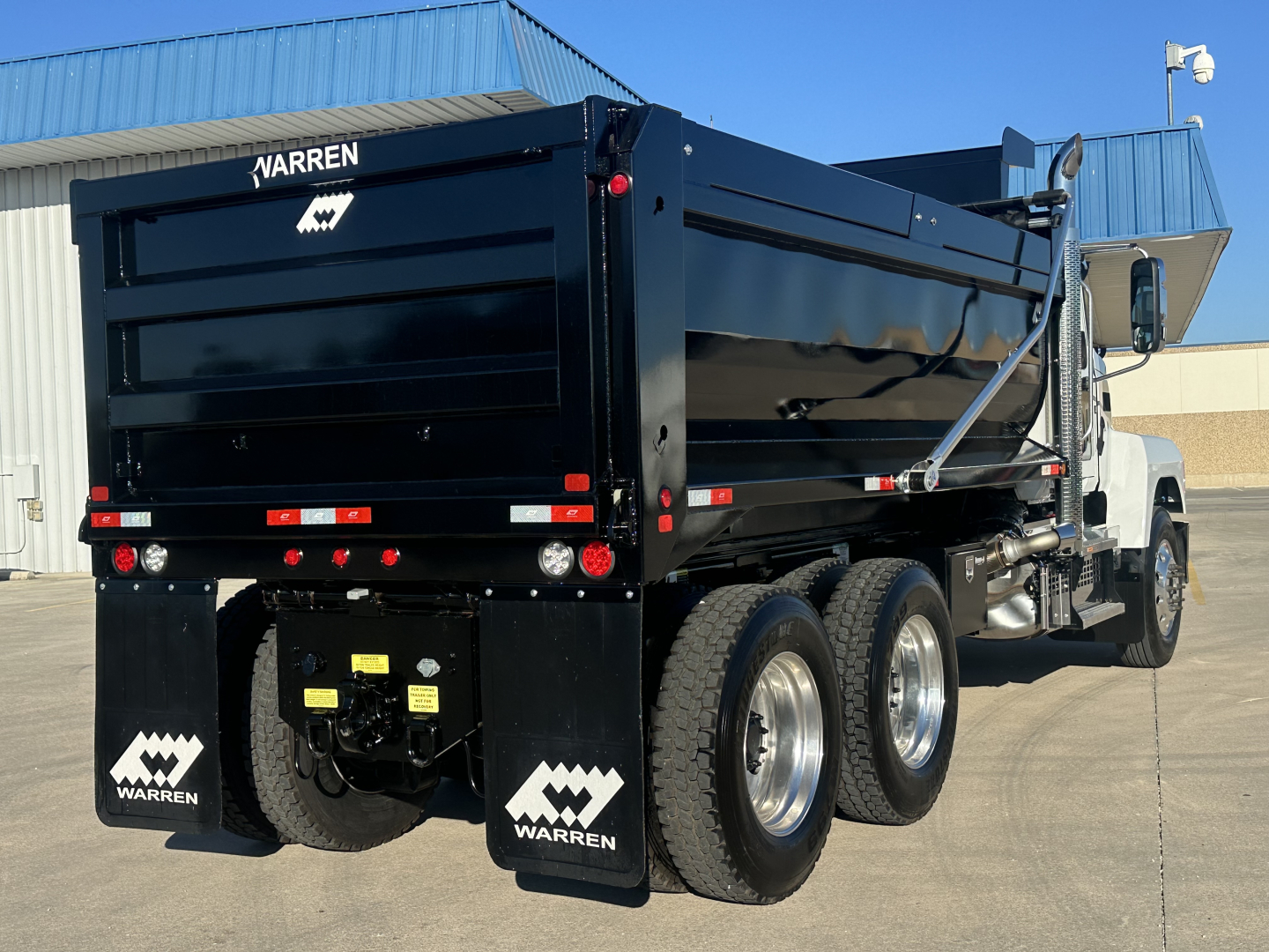 Dump truck rear and side view showing a black Mack PI64 dump truck with Warren logo mud flaps and dual rear axles, bed raised slightly.