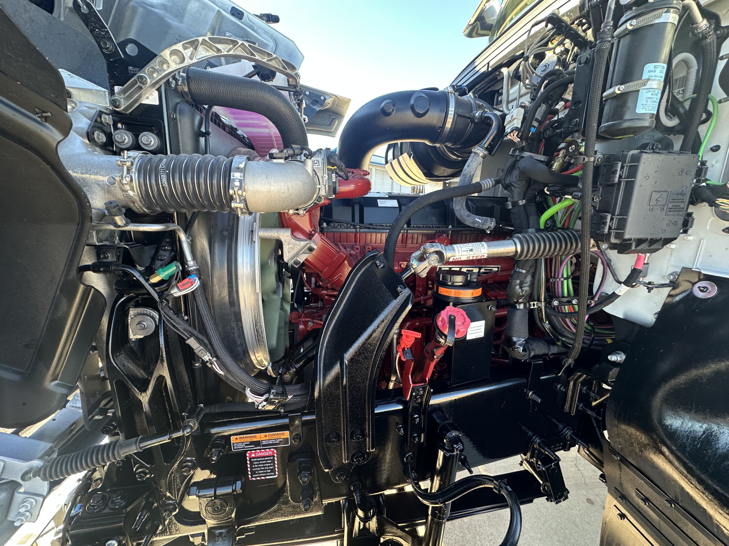 Front engine bay of a 2026 Mack PI64 dump truck showing engine components, hoses, wiring, and red coolant/oil reservoir cap.