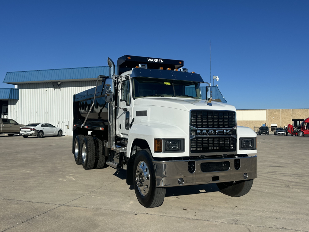 Mack PI64 dump truck in a dealership lot, front-right view, with chrome bumper and grille, large mirrors, and a Warren crane on top of cab.