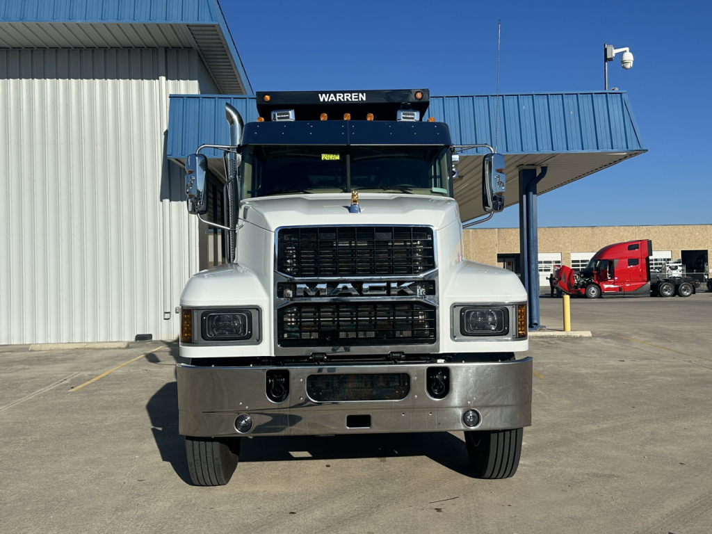 Front view of a white 2026 Mack PI64 dump truck showing grille, headlights, bumper, and 'WARREN' sun visor on top with blue building behind.