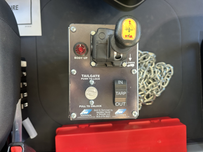Mack PI64 truck controls panel showing body up switch, tailgate lock, and safety lanyard with yellow emergency handle; label text includes IN TARP OUT and warnings.
