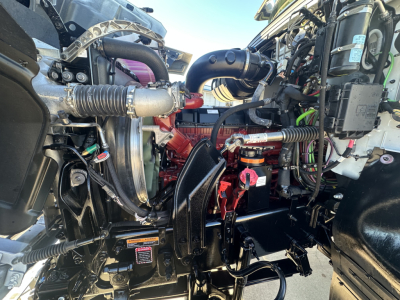 Front engine bay of a 2026 Mack PI64 dump truck showing engine components, hoses, wiring, and red coolant/oil reservoir cap.