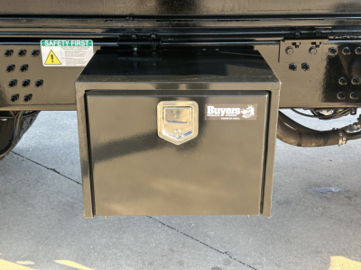 Black rectangular toolbox mounted under the chassis of a Mack PI64 truck showing Buyers aluminum lock box with emblem; safety sticker and frame details visible.