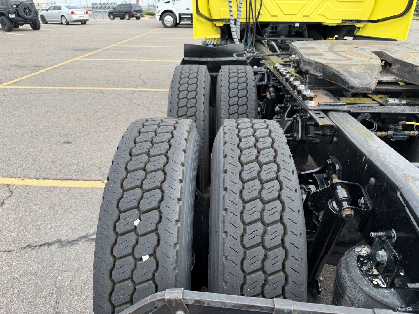 Rear view of a truck chassis showing dual rear tires and part of the cab in yellow; visible components include the suspension, drive axle, and frame.