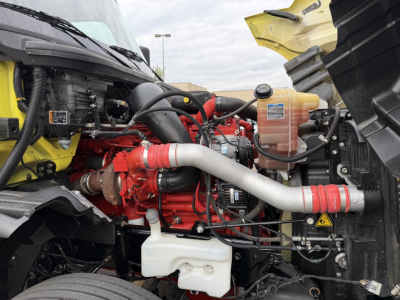 International LT625 engine bay showing Cummins/isx style engine, red engine block with white coolant reservoir and hoses, yellow hood edge, and plastic coolant reservoir with warning labels.