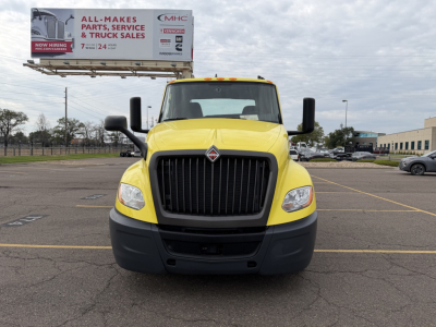 Yellow 2023 International LT625 truck front view in a parking lot with a billboard in the background. Visible text on billboard: ALL-MAKES PARTS, SERVICE & TRUCK SALES. Truck grille and headlights clearly visible.