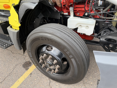 Wheel and front engine area of a 2023 International LT625 conventional daycab showing large front tire, dual tread, wheel hub, brake lug nuts, and engine components with red engine block and white reservoir.