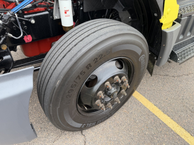 Q: 2023 International LT625 front wheel showing tire size 12R22.5 and hub with lug studs; truck frame and steps visible; part of engine bay and hydraulic lines in background.