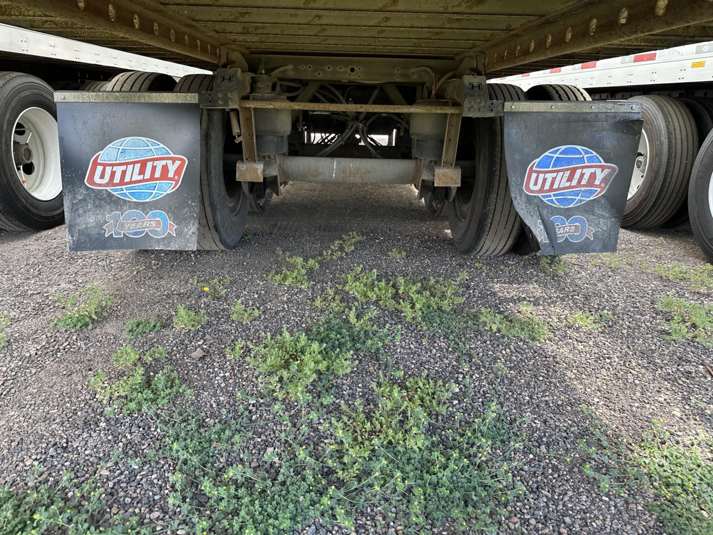 Trailer underside showing two mud flaps with Utility logo and 100-year emblem, frames and axle components, weeds on ground under trailer.