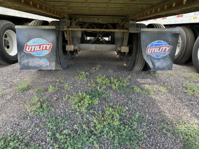 Trailer underside showing two mud flaps with Utility logo and 100-year emblem, frames and axle components, weeds on ground under trailer.