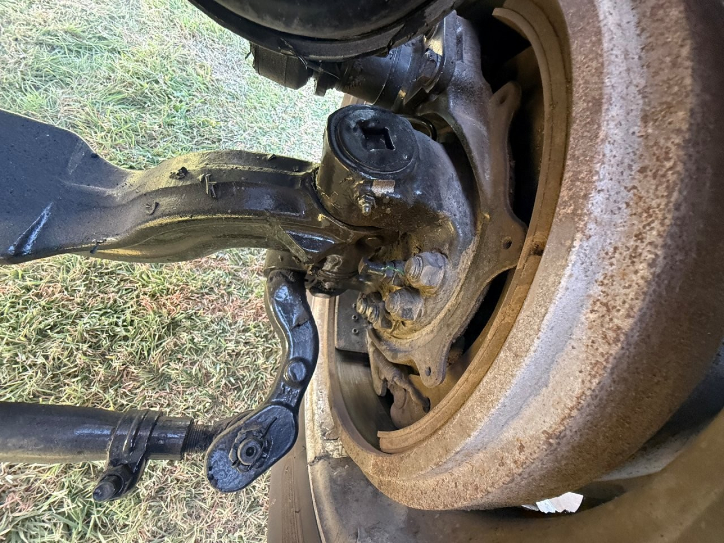 Front axle and suspension of a Mack PI64T showing steering knuckle, coil spring, and brake assembly with exposed wheel hub and drum; dirt and grime on components.