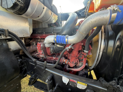 Engine bay of a 2020 Mack PI64T conventional daycab showing red engine block, silver intake pipe, blue silicone coupler, red hoses, and surrounding components.