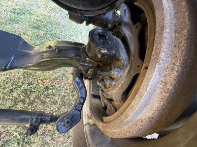 Front axle and suspension of a Mack PI64T showing steering knuckle, coil spring, and brake assembly with exposed wheel hub and drum; dirt and grime on components.