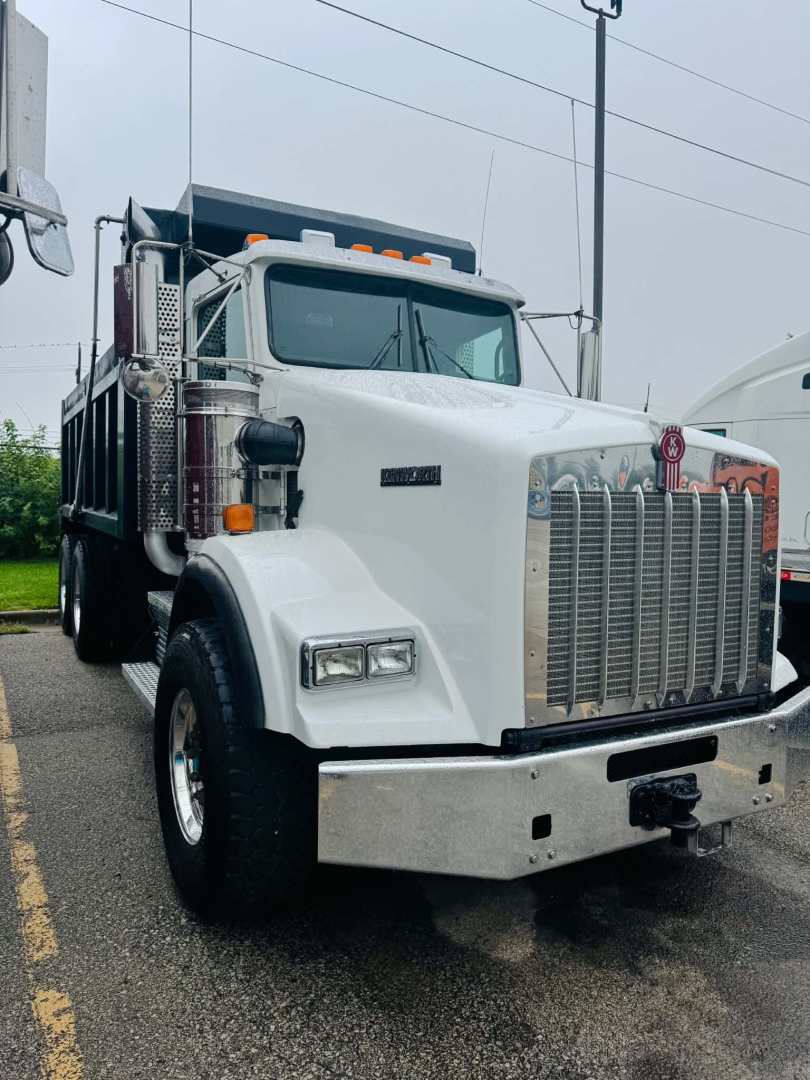 Kenworth T800 dump truck front view with large chrome grille and bumper, white cab, diesel engine bay, visible headlights and side mirrors.