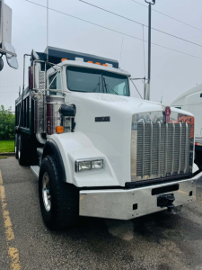 Kenworth T800 dump truck front view with large chrome grille and bumper, white cab, diesel engine bay, visible headlights and side mirrors.