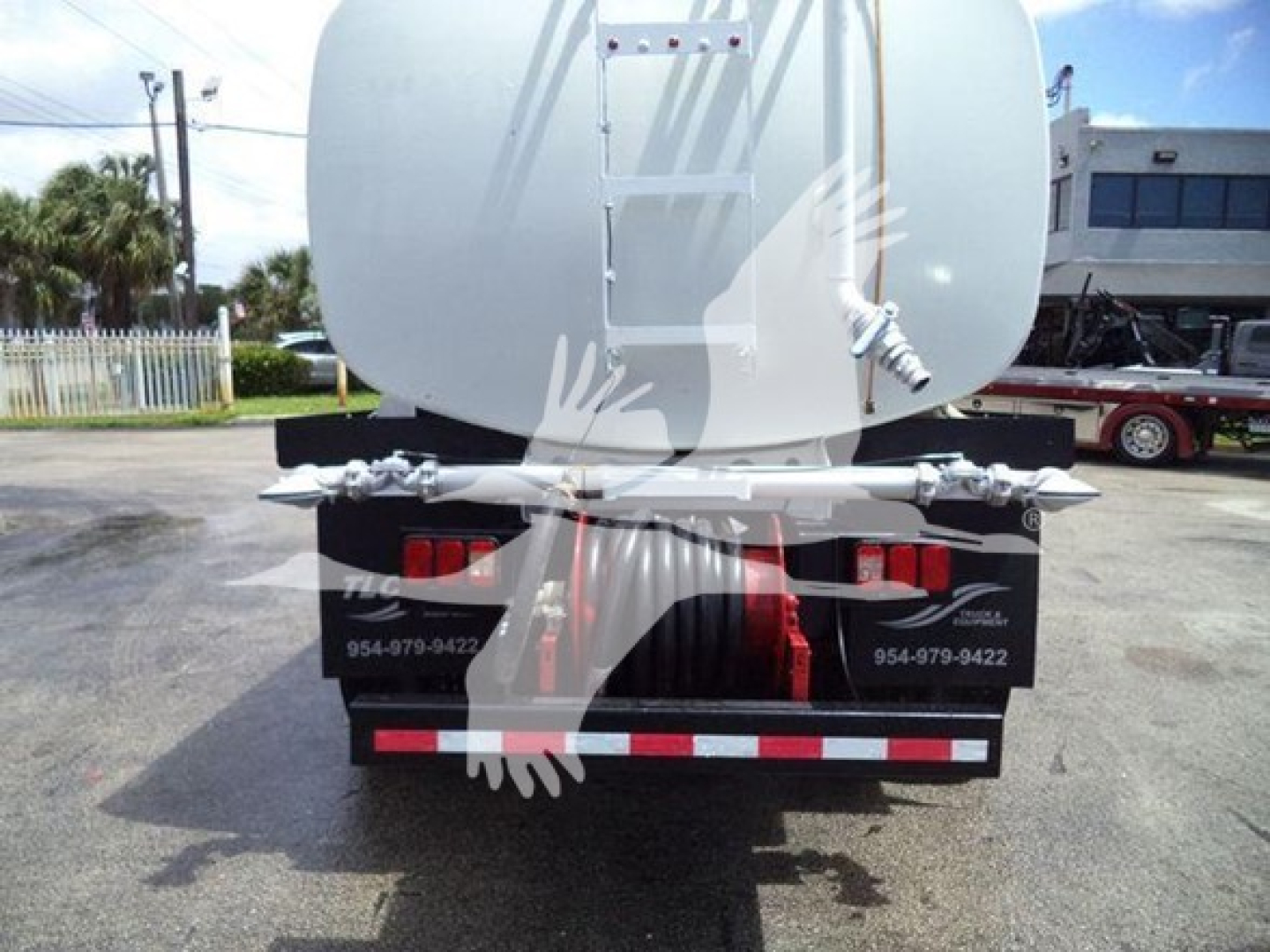 Rear view of a white tanker truck with a rounded tank, dual rear lights, and reflective red/white bumper stripes. Includes contact numbers on black mud flaps and metal ladder on tank.