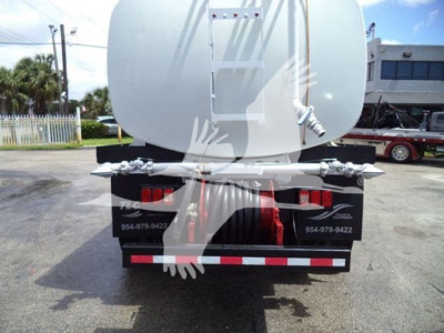 Rear view of a white tanker truck with a rounded tank, dual rear lights, and reflective red/white bumper stripes. Includes contact numbers on black mud flaps and metal ladder on tank.