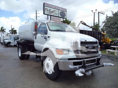 Front-left view of a white 2015 Ford F650 SD gas or diesel truck with a tanker body, showing front bumper, dual headlights, and side step; background includes dealership signage.