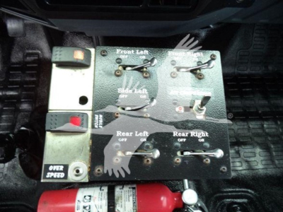 Control panel with multiple toggle switches and labels including Front Left, Front Right, Rear Left, Rear Right, Over Speed; red fire extinguisher handle visible beneath; textured floor mats and pedal nearby in vehicle interior.