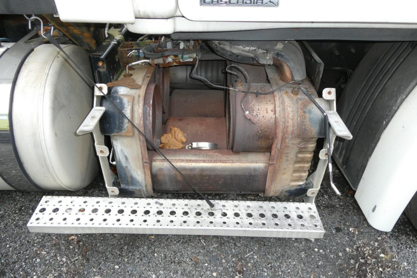 Image of a Freightliner Cascadia 113 from the side showing the undercarriage with a large cylindrical diesel tank and exhaust/muffler area; visible components include a perforated metal step and surrounding frame.