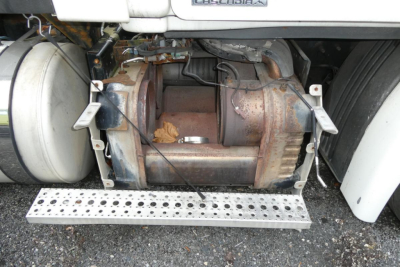 Image of a Freightliner Cascadia 113 from the side showing the undercarriage with a large cylindrical diesel tank and exhaust/muffler area; visible components include a perforated metal step and surrounding frame.