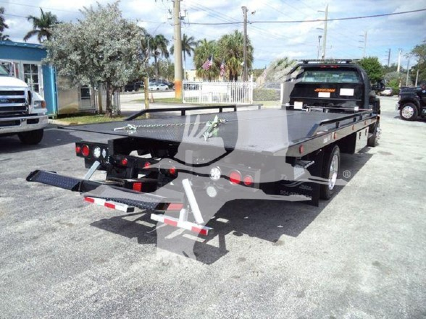 Flatbed tow truck in a dealership lot with rear view showing winch and dual rear wheels.