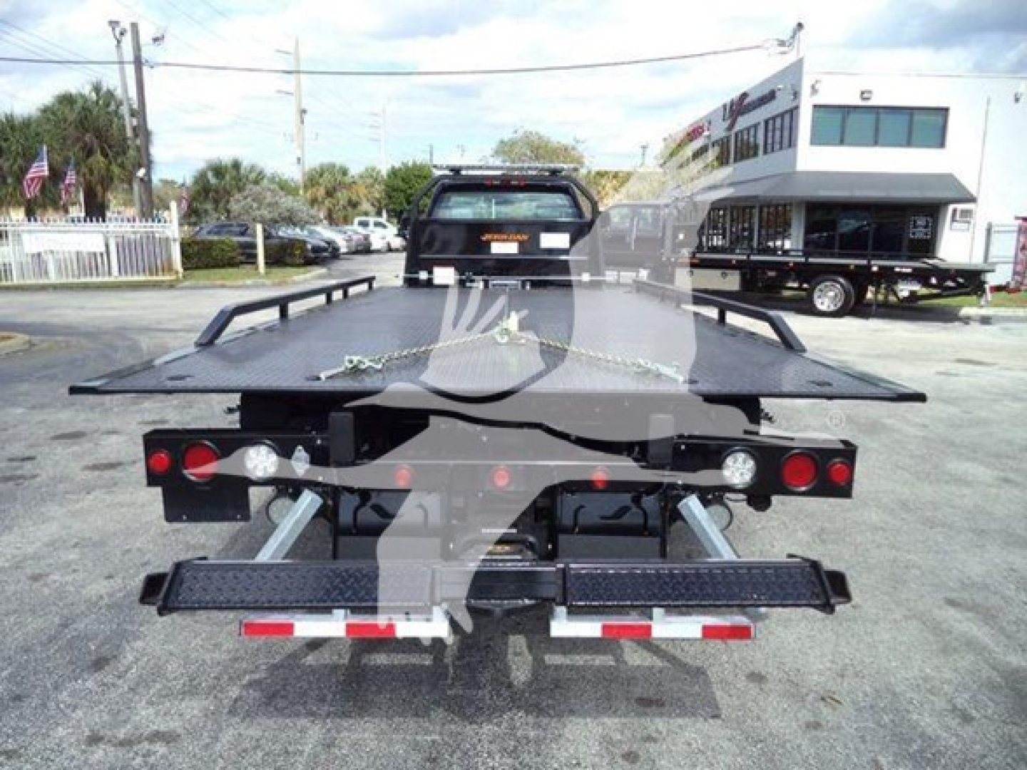 Flatbed tow truck trailer rear view showing dual wheel platforms and rear lights; black metal frame with reflectors and safety rails.