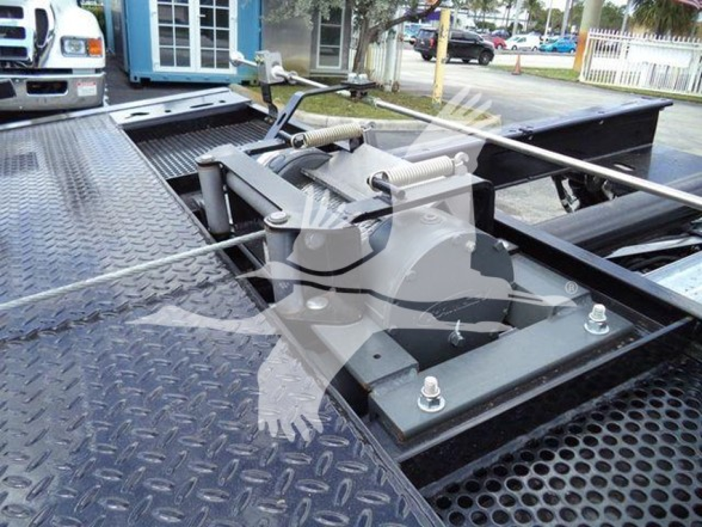 Platform bed with hydraulic system on a rollback tow truck showing winch assembly and metal grid surface.