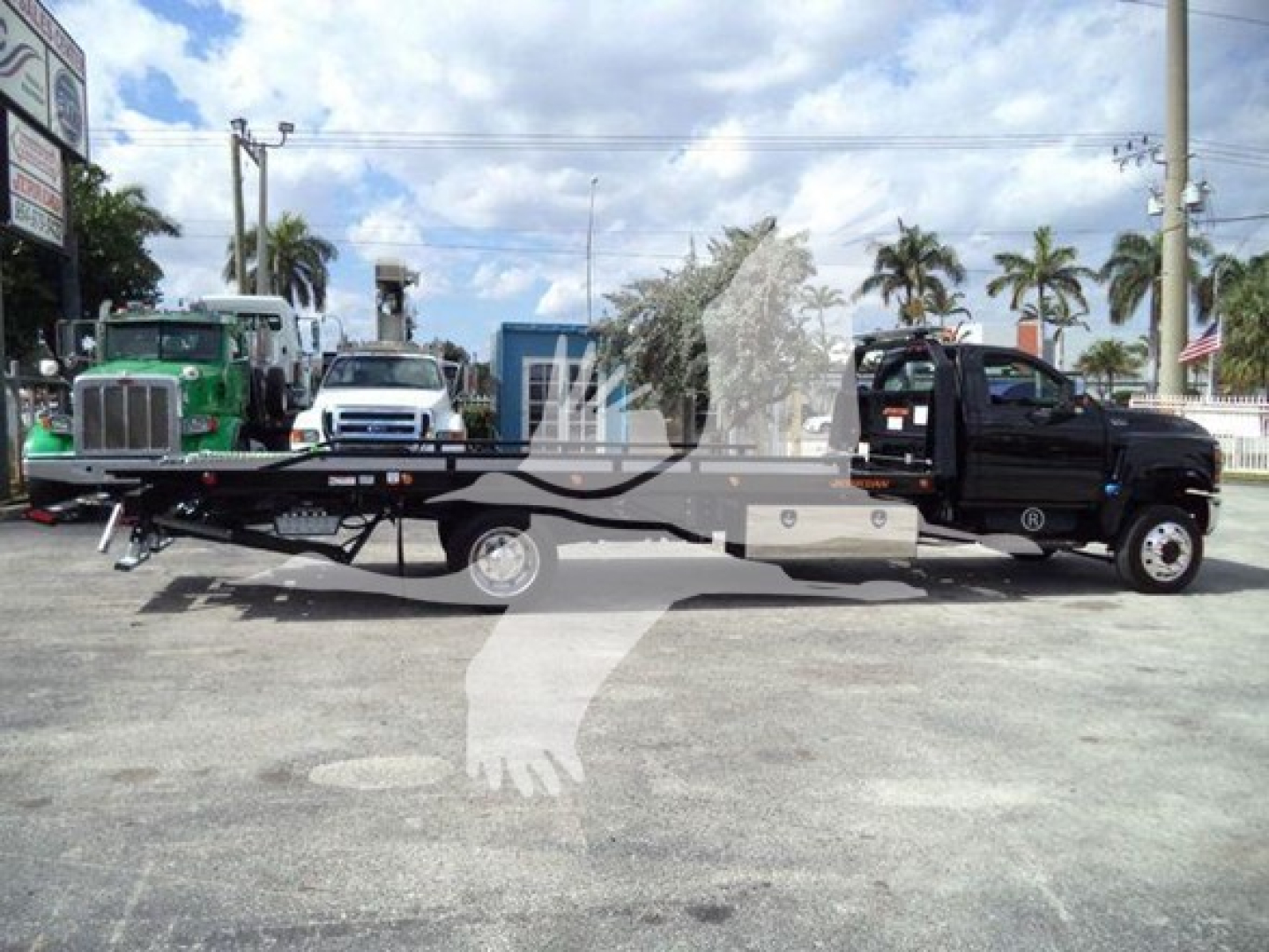 Vehicle side view showing a black rollback tow truck with a flatbed in a truck yard, multiple trucks in the background, palm trees, and clear sky.