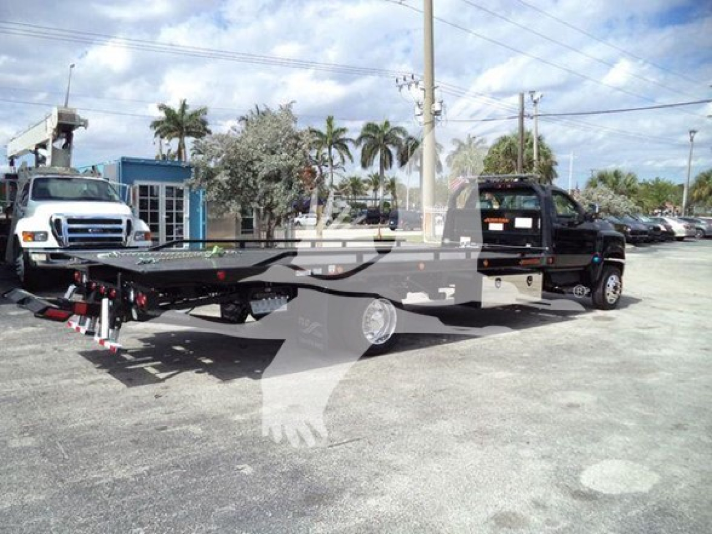 Truck bed rollback tow truck attached to a wrecker with a long flatbed and hydraulic equipment in a dealership lot.