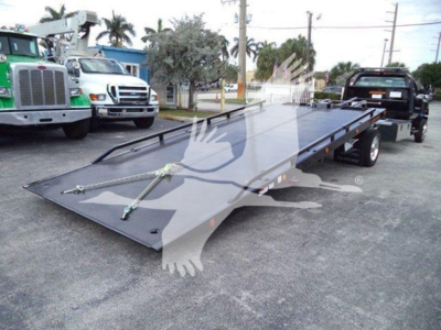 Flatbed tow truck or rollback with a large metallic loading ramp on a trailer, parked in a dealership lot with other trucks in background.