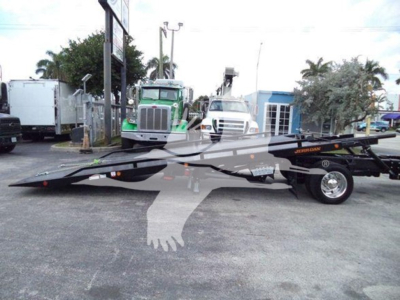 Tractor wrecker rollback bed on a truck with flatbed and wheel. Visibility of a long black tow truck platform and undercarriage with tires; surrounding lot with other trucks.